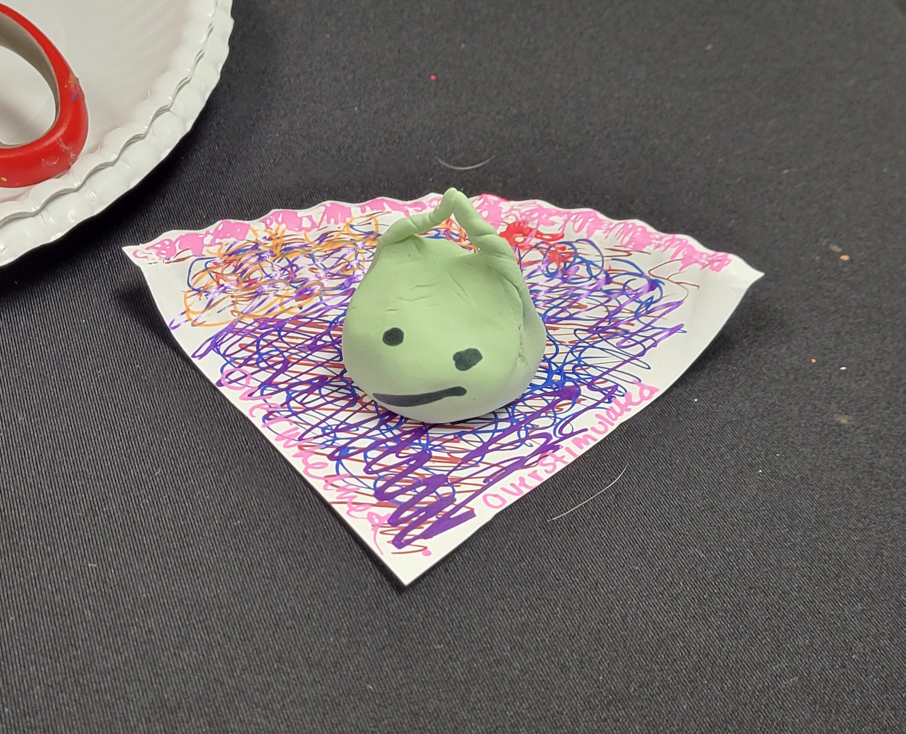 Green alien character made from model magic sitting on a scribbled paper plate. "Overstimulated" is written on the plate.